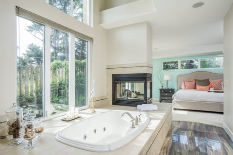 The gorgeous primary suite features a king bed, soaking tub near large windows, and a double-sided fireplace.