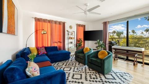 A bright living room featuring two large blue sofas, a TV, and a piano.