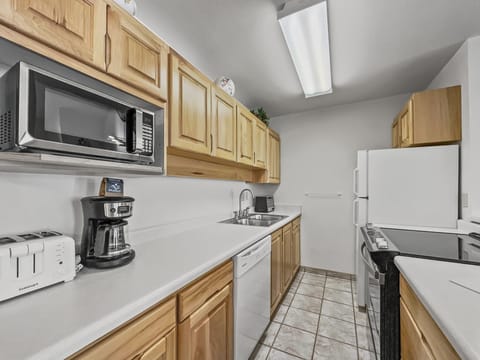 A modern kitchen with wooden cabinets, a microwave, a coffee maker, a toaster, a dishwasher, a refrigerator, a stove, and overhead fluorescent lighting. The countertops are white, and the flooring is tiled.