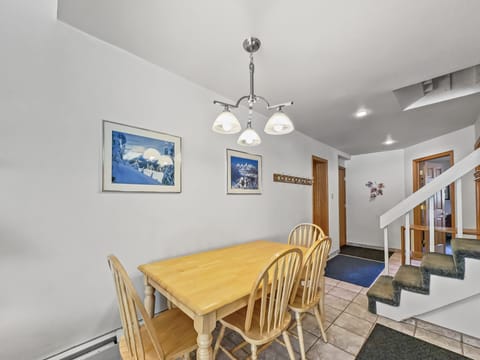 A dining area with a wooden table and four chairs, illuminated by a ceiling light fixture. Two landscape paintings hang on a white wall. A staircase with green carpet and tile flooring is visible.
