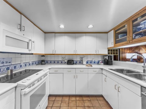A kitchen with white cabinets, a stove, a microwave, a sink, and a coffee maker. The backsplash features blue and white tiling, and the floor is covered with brown tiles.