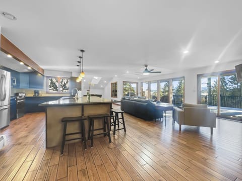 Modern open-concept living area featuring a kitchen with island and bar stools, a dining area, and a spacious living room with large windows and wooden flooring.