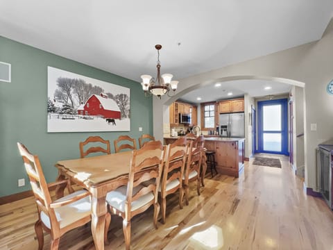 A dining room with a wooden table and eight chairs, a chandelier above, a painting of a red barn on a green wall, and a view into a kitchen with stainless steel appliances.