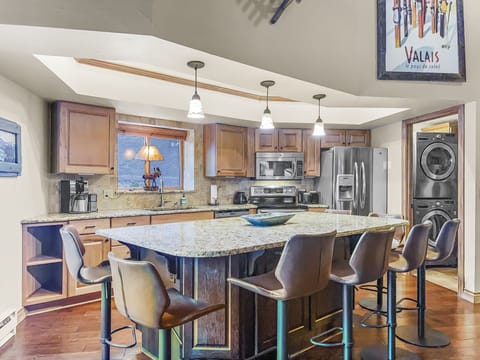 A modern kitchen with granite countertops, stainless steel appliances, wooden cabinets, an island with barstools, and laundry machines in a side nook. A framed poster on the wall reads "Valais.