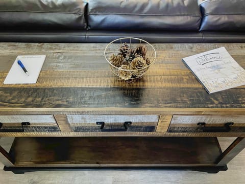 A wooden coffee table with a pen and notepad on the left, a decorative bowl with pinecones in the center, and a "Welcome" brochure on the right. A leather couch is in the background.
