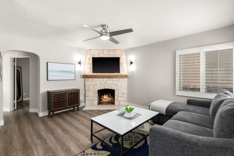 Stylish living room with a stone fireplace, wooden mantel, and mounted TV. Modern gray sectional sits beside a marble coffee table. Ceiling fan enhances comfort and airflow.