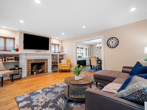 Bright and cozy living room with a comfy sectional 🛋️, floral rug 🌸, and a charming fireplace 🔥. Open layout flows into a stylish dining area — perfect for relaxing or entertaining 🍷✨.