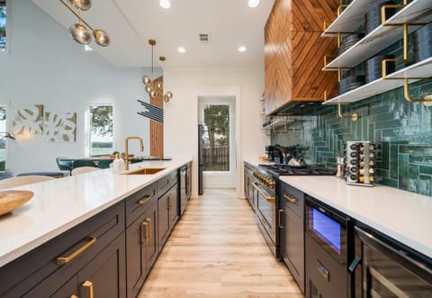 A Stylish Kitchen Space Featuring Beautiful Cabinetry, Elegant Gold Accents, White Countertops, And Floating Wooden Shelves, With Natural Light Streaming In Through A Door At The End Of A Gall