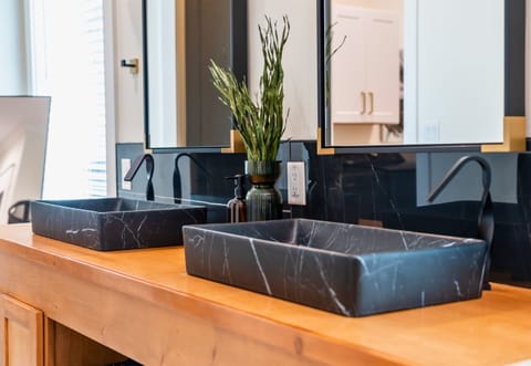 A Close-up View Of A Marbled Vessel Sinks Paired With Matte Black Faucets On A Wooden Countertop, Complemented By A Simple Greenery Arrangement That Adds A Touch Of Organic Freshness To The Modern, Clean Design.
