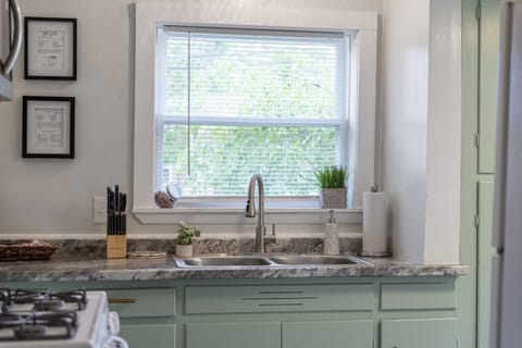 Compact galley kitchen features white appliances including a fridge and gas range under a microwave, paired with muted green cabinets and a double sink beneath a window. Bright and functional space 🍳🌿🔑