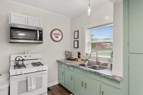 Compact galley kitchen features white appliances including a fridge and gas range under a microwave, paired with muted green cabinets and a double sink beneath a window. Bright and functional space 🍳🌿🔑