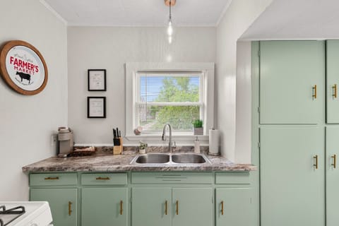 Compact galley kitchen features white appliances including a fridge and gas range under a microwave, paired with muted green cabinets and a double sink beneath a window. Bright and functional space 🍳🌿🔑