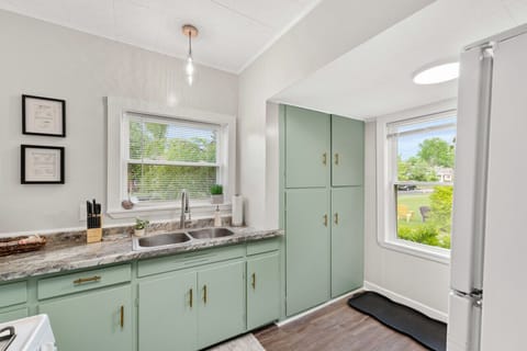 Compact galley kitchen features white appliances including a fridge and gas range under a microwave, paired with muted green cabinets and a double sink beneath a window. Bright and functional space 🍳🌿🔑