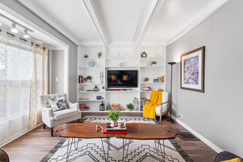 Bright living space with a plush sofa and accent chairs arranged around a live-edge coffee table on a patterned rug. Built-in white shelves flank a wall-mounted TV, while sheer curtains filter soft natural light through large windows. 🛋️📺🌿