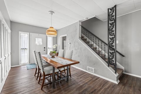 Bright dining area showcases a live-edge wooden table with a lace runner, surrounded by white upholstered chairs and topped with a bowl of green apples. A woven pendant light hangs above, while a row of white louvered closet doors lines the gray wall. Sun