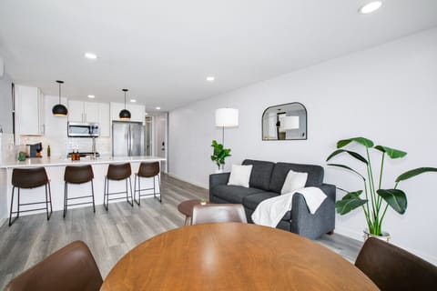 A spacious and modern open-plan living area featuring a sleek kitchen with a large island, a cozy gray sofa, and a stylish dining table. The room is adorned with contemporary decor and indoor plants, creating a welcoming and comfortable space for both rel