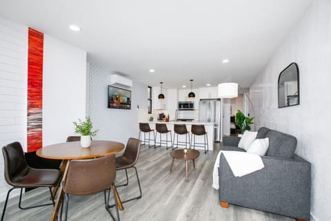 A chic and inviting open-concept living space featuring a modern kitchen with a large island and bar seating, a cozy gray sofa, and a stylish dining area. The room is enhanced by contemporary artwork and decor, creating a perfect setting for both relaxati