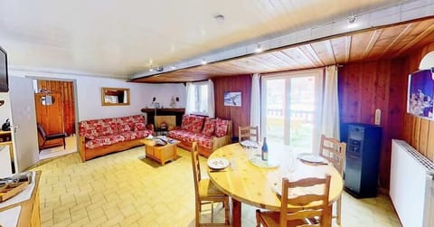 The spacious living area features a comfortable sofa with colorful patterned cushions, a wooden coffee table, and a dining table with chairs set for four. The large window provides a view of the outdoor terrace and surrounding natural landscape. The woode