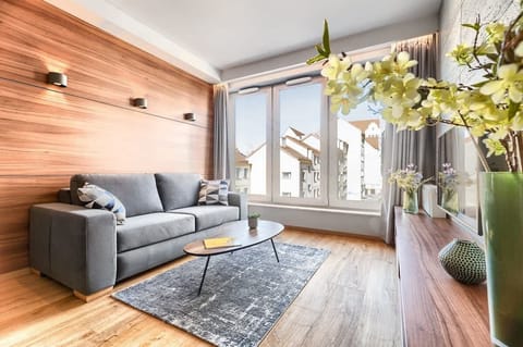 Bright living room with a grey sofa, wooden accents, large window, and a vase with yellow flowers.