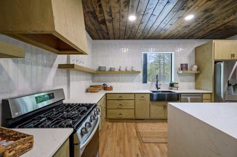 Stylish kitchen with wood cabinetry, stainless steel appliances, farmhouse sink, open shelving, and natural light through a large window with forest views.