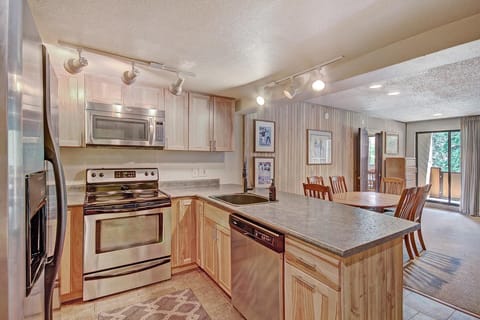 Modern kitchen with stainless steel appliances, wooden cabinetry, and a countertop that extends into a dining area with a wooden table and chairs. A balcony is visible through sliding glass doors.