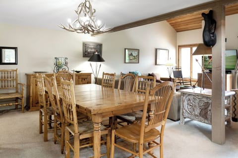 A rustic dining room features a wooden table with eight chairs, an antler chandelier, and assorted decor. The adjacent living area includes a sofa, armchairs, and a TV.