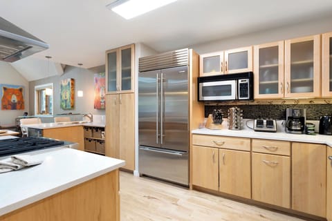 A modern kitchen with light wooden cabinets, stainless steel appliances, including a refrigerator and microwave, and various small kitchen appliances on the countertop. Art pieces hang on the walls.