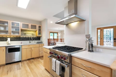 Modern kitchen with light wood cabinetry, stainless steel appliances, a gas cooktop with range hood, and an island. The room is well-lit and opens to a dining area with wooden furniture.