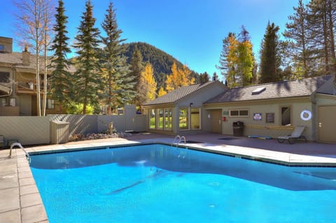 Outdoor swimming pool with clear blue water, surrounded by trees and a building with large windows. A lifebuoy is mounted on the wall, and mountains are visible in the background.