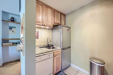 Compact kitchen with wooden cabinets, granite countertops, stainless steel sink, and fridge. Dishwasher below the counter. White tile floor and a silver trash can in the corner.
