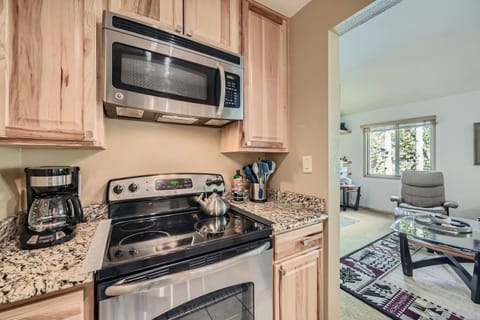 Kitchen with a microwave, electric stove with a kettle, coffee maker on a granite countertop; doorway leading to a living room with a chair and glass coffee table.
