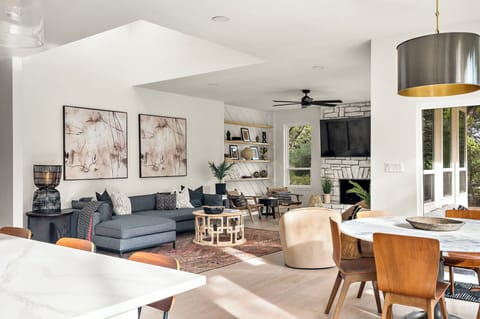 Elegant open-concept living and dining area featuring a cozy gray sectional, abstract wall art, a stone fireplace with a mounted TV, built-in shelves, and a stylish round dining table under a modern brass pendant light. 