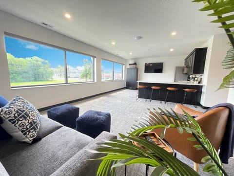 A spacious lounge area bathed in natural light from wall-to-wall windows, featuring a cozy sectional, stylish poufs, and modern seating. Perfect for relaxing or entertaining with scenic views and seamless access to the kitchen and bar.