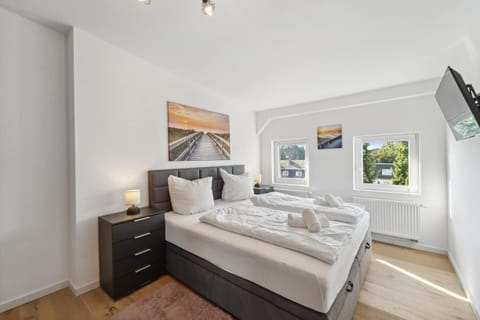 Das Schlafzimmer ist hell und freundlich eingerichtet, bietet ein großes Boxspringbett mit frischer Bettwäsche, zwei Nachttische mit Lampen, Wandbilder mit Strandmotiv sowie einen an der Wand montierten Fernseher.