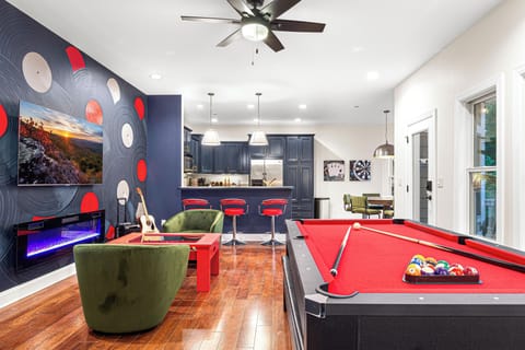 A vibrant entertainment space featuring a red-felt pool table, a cozy seating area with green velvet chairs, and a modern electric fireplace beneath a mounted TV. The stylish bar area, adorned with navy blue cabinetry and red barstools, seamlessly connects to a game corner with a dartboard and poker artwork, creating the ultimate game room experience.
