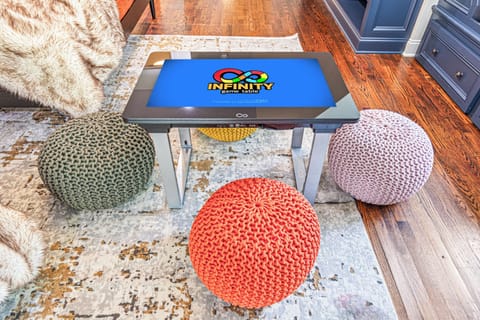 A cozy and interactive gaming nook featuring an Infinity Game Table, surrounded by colorful knitted poufs for comfortable seating. The setting combines modern technology with a warm, inviting ambiance, complete with a soft area rug, faux fur accents, and rich hardwood flooring, creating the perfect space for social gaming and relaxation.