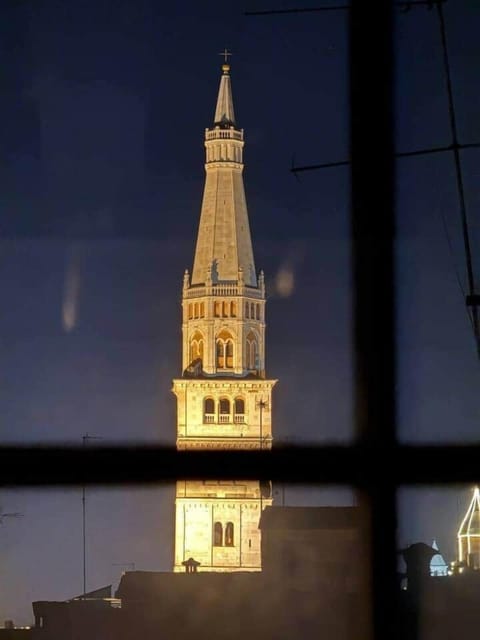 Sdraiato sul letto ammiri la Torre della Ghirlandina incorniciata dalla finestra: Modena ti entra nel cuore già al risveglio. Travi a vista, luce naturale e silenzio creano un rifugio sospeso tra charme e tranquillità.