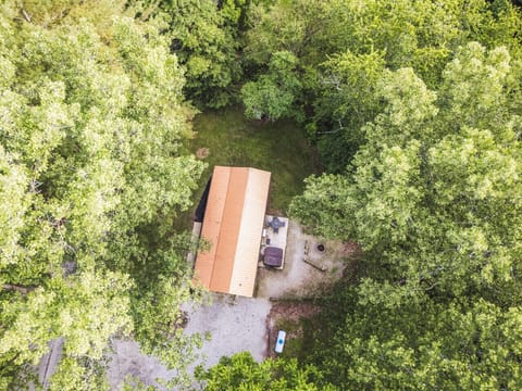 Aerial view of a cabin surrounded by lush green trees, featuring a gravel driveway, outdoor patio with furniture, and a fire pit area — perfect for a serene nature getaway 🌳🏡🔥