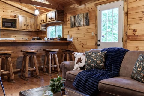 Cozy cabin kitchen and living area with rustic wooden bar stools, plush sofa with nature-themed pillows and a soft navy throw. Inviting space with natural wood finishes and warm lighting for relaxing and socializing 🪵🍂🛋️