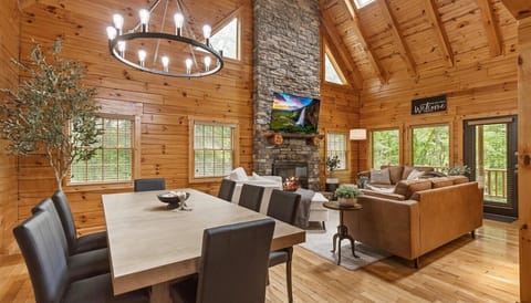 Beautiful and cozy interior of our modern cabin. The space features an open-concept living and dining area with an abundance of natural wood. The living area boasts a grand stone fireplace that extends up to the vaulted ceiling with modern styling.