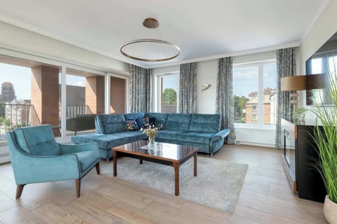 A spacious living room with a large round chandelier, a teal sofa, and a dining area in the background.