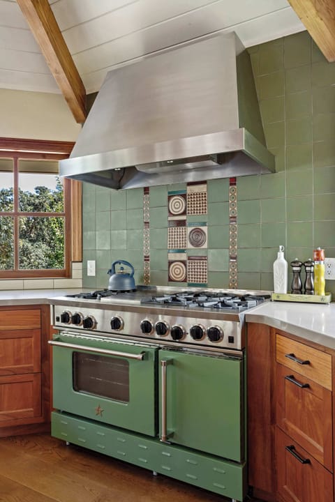 Unique kitchen corner with a vintage green range and oven below a large stainless steel hood, set against a green tiled backsplash and warm wooden cabinets.