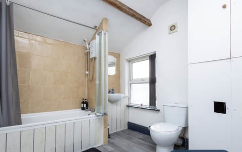 A clean, modern bathroom with a bathtub featuring a wooden panel, beige tiled wall, and glass shower screen. It includes a white toilet, sink, and storage cabinets, with grey flooring and a small window for natural light.