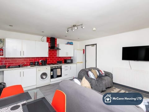 Modern open-plan kitchen and living area with white cabinets, bold red tiled backsplash, and integrated appliances. Comfortable grey sofas face a wall-mounted TV. Red dining chairs add a pop of colour to the sleek, bright space.