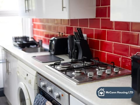 A modern kitchen with white cabinets, a washing machine, and a gas stove. The backsplash features glossy red tiles, adding a vibrant touch. Black utensils and a kettle sit neatly on the countertop for convenience and style.