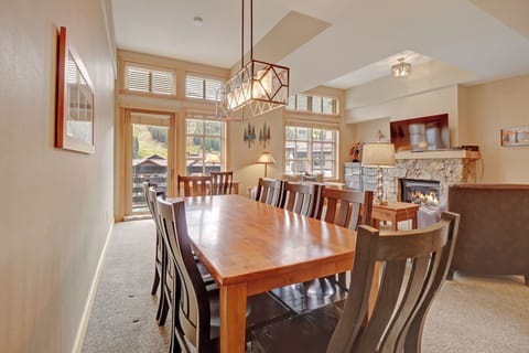 Spacious dining area featuring a large wooden table for eight, illuminated by a contemporary chandelier. Enjoy mountain views through expansive windows and the cozy ambiance of the nearby stone fireplace.
