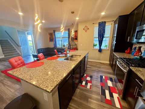 Open kitchen with a long island & bar. The island has a sink and plenty of counter space. Cabinets & modern appliances line the wall, & windows bring in natural light for a cozy space to cook.