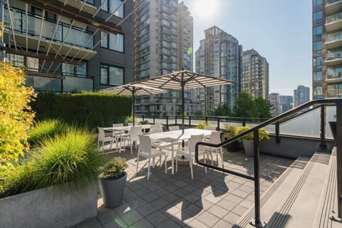 The rooftop terrace offers a serene escape with modern white furniture shaded by striped umbrellas, set against a backdrop of towering glass skyscrapers under the bright sun.