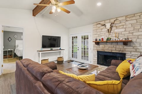 Cozy living room anchored by a brown leather sectional, reclaimed wood coffee table, yellow accent pillows, and a stone fireplace with a rustic mantel — a warm, inviting Texas-style retreat with French doors that flood the space with natural light.