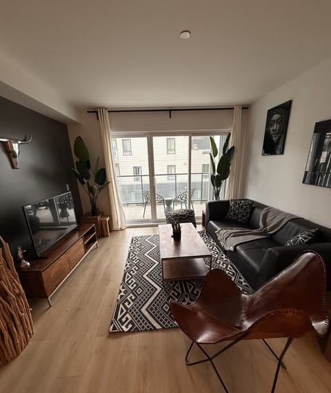 Relax in a beautifully decorated, open living space featuring a deep leather sofa, iconic butterfly chair, geometric area rug, and flat-screen TV—all framed by natural light and balcony access.